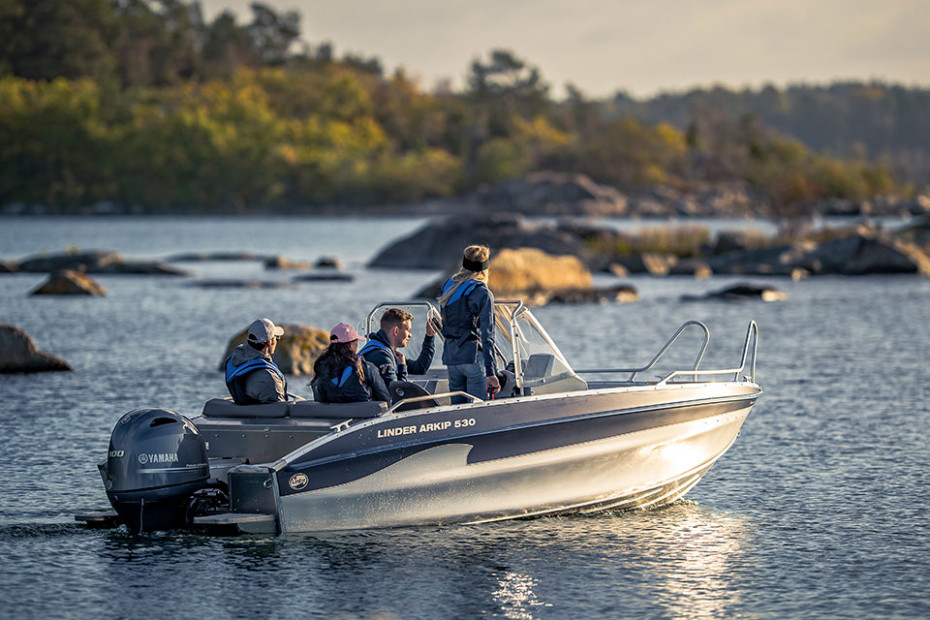 Idyllisk bild av svensk kust, i kvällssol. Fyra unga personer i en aluminiumbåt från Linder.