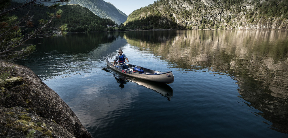 En man paddlar en Inkas kanot på ett stilla vatten omgivet av skogsbeklädda berg.