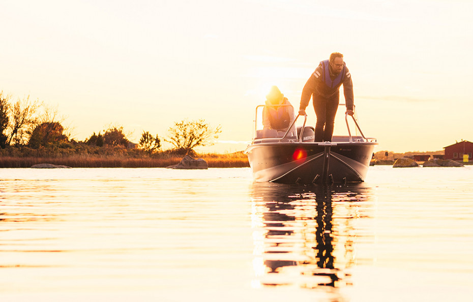 En man står i fören på en Linderbåt och gör sig redo att hoppa iland. Det är kvällssol i bilden och vattnet är stilla.
