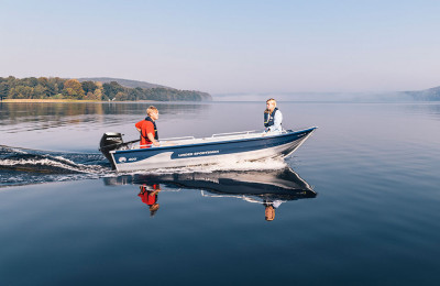 En spegelblank sjö en solig dag. Två personer kör Linderbåten Sportsman 400.