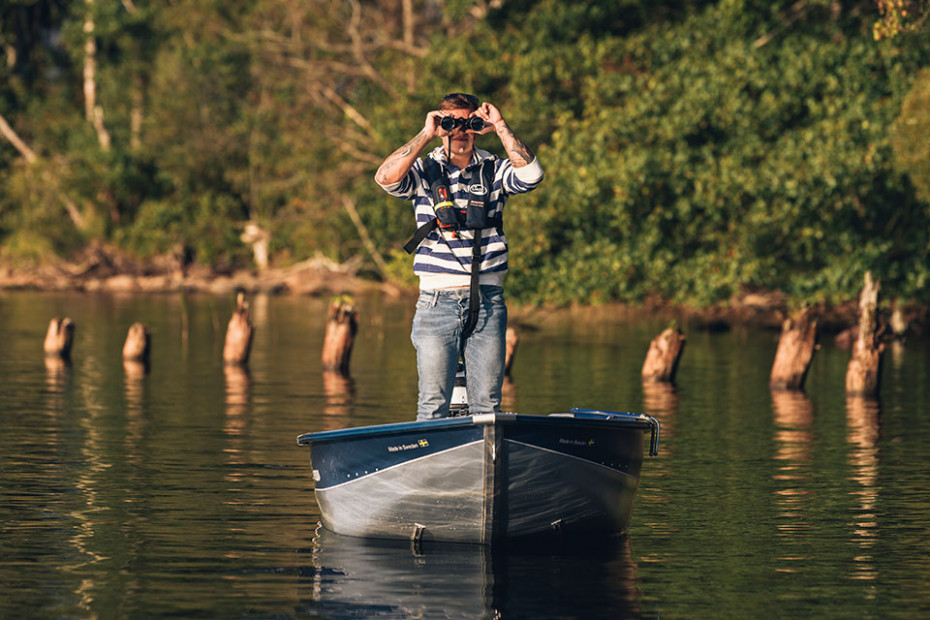 Bilden tagen framifrån. Man står i aluminiumbåt och tittar i kikare. I bakgrunden vatten och skog.