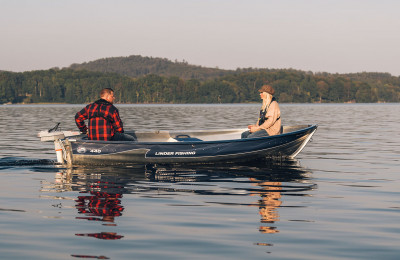 Bästsäljande Fishing 440 förs fram sommartid på ett stilla vatten av en man i rödrutig jacka. En person sitter i fören.