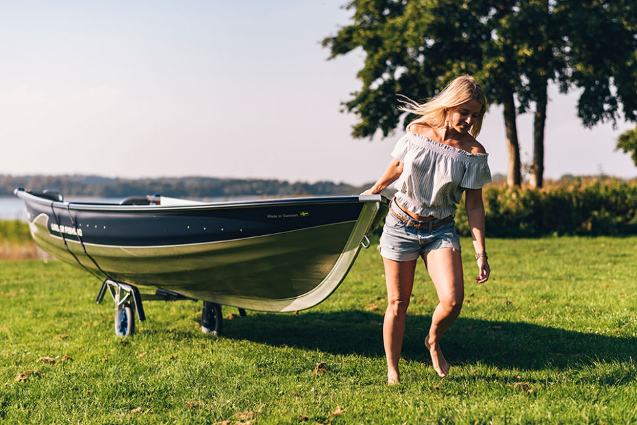 En vacker kvinna drar en Linderbåt på en båtvagn över en gräsmatta i solen.