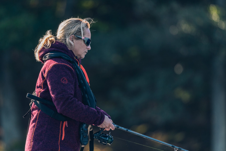Halvkroppsbild i profil på sportfiskande kvinna som är ute på sjön. Hon har vinröd jacka och i bakgrunden är det skog..
