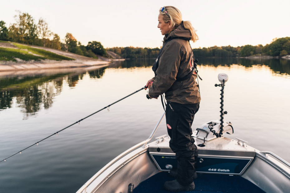 Kvinna sportfiskar från sin fiskebåt på stilla vatten en tidig morgon eller kväll. Klippstränder i bakgrunden.